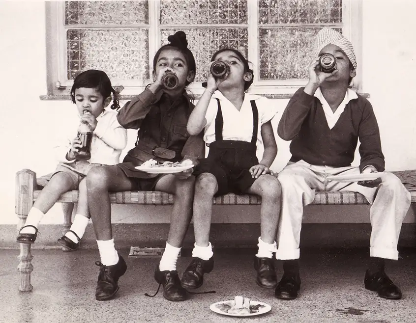 Photo captured in 1964 in Arusha, Kenya, featuring four children sitting on a bench and drinking from glass bottles.